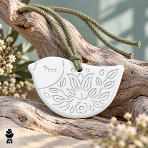 May include: A white ceramic bird ornament featuring an embossed floral pattern and a closed eye design. The bird is suspended by a thick olive green braided cord, showcasing a smooth matte finish and intricate carved details on its surface.