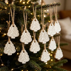 May include: A collection of white, flat, clay-like Christmas tree ornaments hanging from jute twine strings. Each ornament features a detailed, embossed swirl and leaf pattern on the surface, shaped like a stylized evergreen tree.