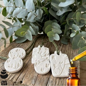 May include: White ceramic essential oil diffusers with embossed botanical designs, hanging on green cords. Eucalyptus leaves and a small amber bottle with a dropper are in the background. The diffusers are on a wooden surface.