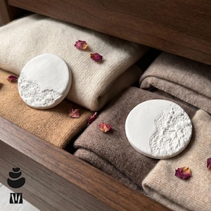 May include: Two circular white clay discs with textured, lace-like relief patterns on one side, resting on folded beige and brown knit fabric. Several small, dried dark pink rose petals are scattered across the fabric surfaces.