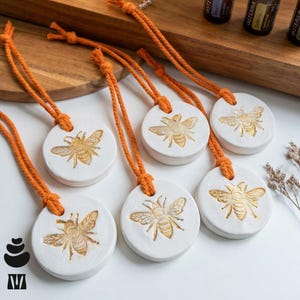 May include: White ceramic essential oil diffusers featuring a gold bee design, suspended by orange cords. The circular diffusers are displayed on a white surface, with a wooden backdrop and essential oil bottles in view.