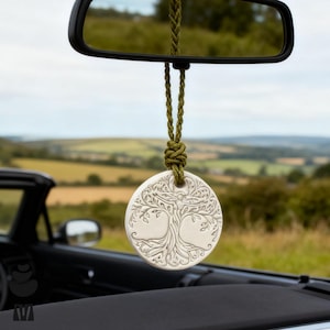 May include: A white, circular car air freshener featuring a detailed tree of life design, suspended from a green braided cord. The air freshener hangs from a car's rearview mirror, with a countryside view in the background.