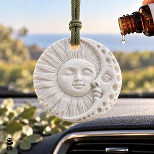 May include: A circular white ceramic hanging ornament featuring a relief design of a sun and a crescent moon with facial features. The sun has radiating rays and the moon has crater textures. It hangs from a green cord, with an amber oil dropper.
