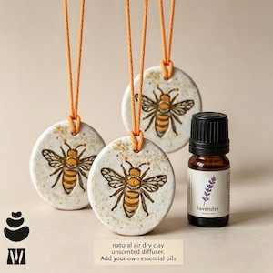 May include: Three round, speckled white clay diffusers featuring a hand-painted bee illustration, each suspended by an orange cord. Beside them sits a small amber glass bottle with a black cap and a white label reading lavender.