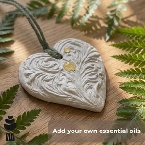 May include: A heart-shaped, off-white essential oil diffuser with an embossed floral design. It has a green cord for hanging and is adorned with drops of oil. The text "Add your own essential oils" is at the bottom.