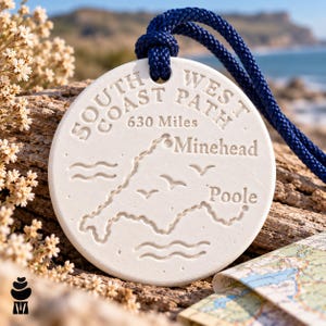 May include: A circular white ceramic pendant featuring the embossed text SOUTH WEST COAST PATH, 630 Miles, Minehead, and Poole. The design includes a map of the trail, wave icons, and birds, suspended by a thick dark blue braided rope cord.