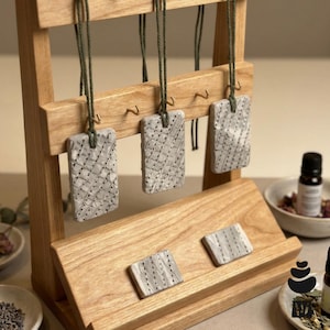 May include: A light-coloured wooden display stand holds three rectangular, textured grey clay pendants hanging from green cords, with two additional matching rectangular clay pieces resting on the lower slanted wooden shelf of the stand.