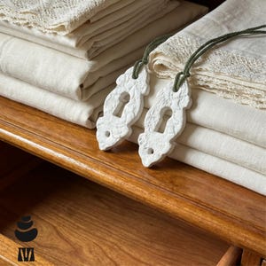 May include: Two white, textured, decorative keyhole-shaped ornaments made of a matte material, each suspended by a thin dark green cord. These ornaments are resting on a stack of folded off-white linen fabric featuring delicate lace trim edges.