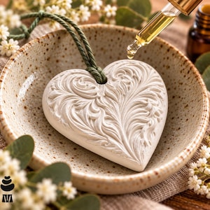 Peut inclure: Un pendentif diffuseur en céramique blanche en forme de cœur avec un motif de volutes en relief, suspendu par un cordon vert. Un compte-gouttes en verre libère une goutte de liquide doré sur la surface texturée du cœur posé dans un bol moucheté.