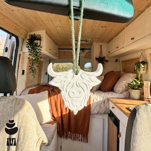 May include: White Highland cow-shaped car air freshener suspended from a green string. The air freshener is in a van with a wooden interior, a bed, and plants. The van has a comfortable, homely atmosphere.