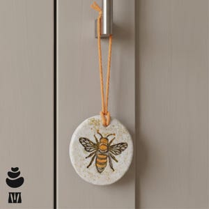 May include: A circular ceramic ornament with a bee design, suspended by an orange string. The bee is depicted with black and orange stripes. The ornament is attached to a silver metal handle.