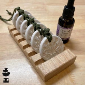 May include: A set of six white, circular aromatherapy stones with a floral design, connected by a green cord, resting in a wooden holder. A bottle of lavender essential oil is next to the holder. The stones are approximately 5 cm in diameter.