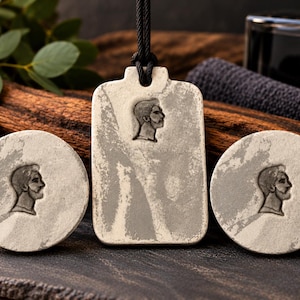 May include: A set of three light grey stone-like accessories featuring a relief profile of a person with a beard. The set includes two circular pieces and one rectangular pendant suspended by a black cord, all displaying a textured, weathered finish.