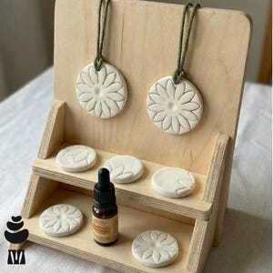 May include: A light wooden display stand features white ceramic daisy-shaped air fresheners and essential oil. Two daisy-shaped air fresheners hang from the top, with others on the shelves. A small brown essential oil bottle is also present.