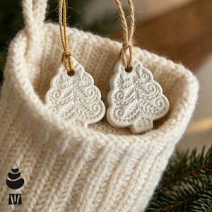 May include: Two white ceramic Christmas tree ornaments with intricate embossed swirl and leaf patterns, each hanging from a tan jute string, placed inside a cream-coloured knitted stocking with a ribbed texture and a loop at the top.