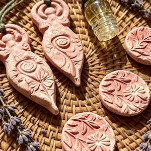 May include: Two terracotta-coloured clay pendants shaped like stylised figures with spiral and floral carvings, each on a green cord, alongside three round clay discs with floral relief patterns, a small glass dropper bottle, and sprigs of dried lavender.