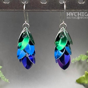 May include: A pair of silver earrings with blue, green, and purple scales. The earrings are hanging from a metal rack. The text "MYCHICA METALWORKS" is visible in the background.