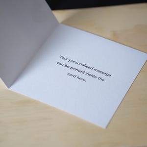 Op de afbeelding: Een open witte wenskaart met de tekst: "Uw gepersonaliseerde bericht kan hier in de kaart worden afgedrukt." De kaart ligt op een lichtbruin houten oppervlak.