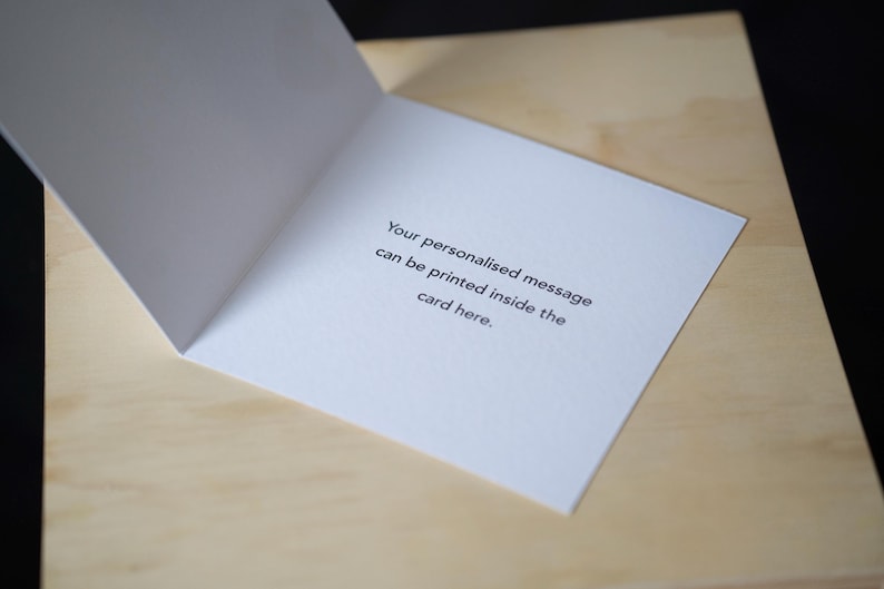 Op de afbeelding: Een open witte wenskaart met de tekst: "Uw gepersonaliseerde bericht kan hier in de kaart worden afgedrukt." De kaart ligt op een lichtbruin houten oppervlak.