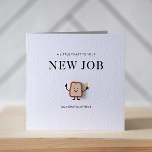 May include: White greeting card with the text "A LITTLE TOAST TO YOUR NEW JOB" in black. A cartoon toast character with a smiling face and a champagne glass is featured below the text. The word "CONGRATULATIONS" is printed at the bottom.