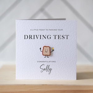 Op de afbeelding: Witte wenskaart met de tekst "A LITTLE TOAST TO PASSING YOUR DRIVING TEST". Een lachend toastkarakter houdt een drankje vast. De kaart zegt "Congratulations Sally".