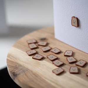 Op de afbeelding: Een houten snijplank met verschillende kleine, bruine, lachende toastvormige magneten verspreid over de plank. E&eacute;n magneet is bevestigd aan een witte kaart. De magneten hebben een lichtbruin midden en een donkerdere bruine rand.