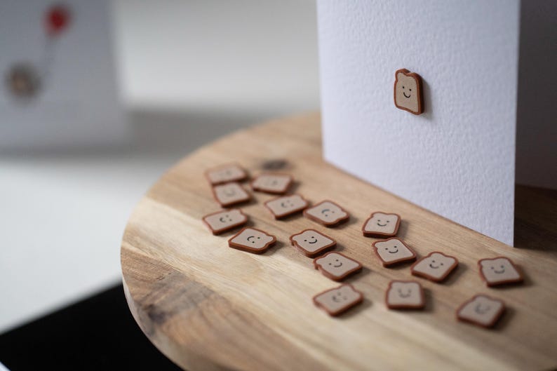 Op de afbeelding: Een houten snijplank met verschillende kleine, bruine, lachende toastvormige magneten verspreid over de plank. E&eacute;n magneet is bevestigd aan een witte kaart. De magneten hebben een lichtbruin midden en een donkerdere bruine rand.
