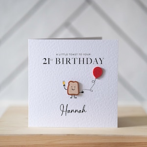 May include: A white birthday card with the text "A LITTLE TOAST TO YOUR 21ST BIRTHDAY" and the name "Hannah". The card features a cartoon toast character holding a drink and a red balloon.
