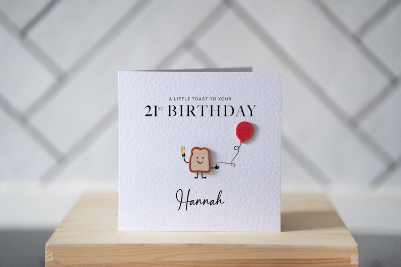May include: A white birthday card with the text "A LITTLE TOAST TO YOUR 21ST BIRTHDAY" and the name "Hannah". The card features a cartoon toast character holding a drink and a red balloon.