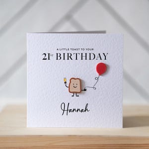 May include: White birthday card with the text "A little toast to your 21st Birthday" and the name "Hannah". The card features a toast character holding a champagne glass and a red balloon.