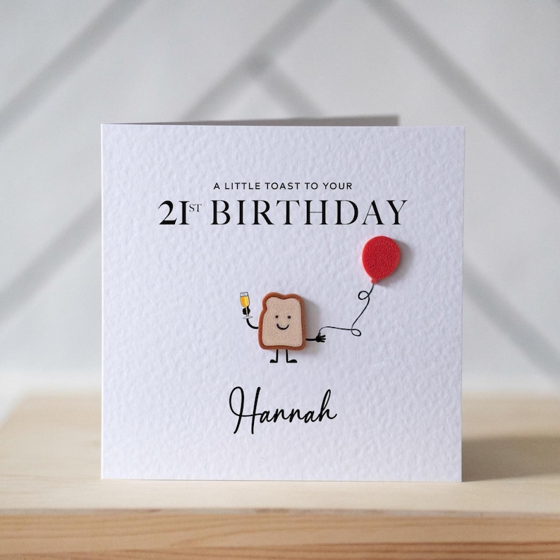 May include: White birthday card with the text "A little toast to your 21st Birthday" and the name "Hannah". The card features a toast character holding a champagne glass and a red balloon.