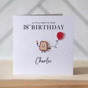 May include: A white square greeting card with the text A LITTLE TOAST TO YOUR 18TH BIRTHDAY and the name Charlie. The design features a cartoon slice of toast with a smiling face, holding a champagne glass and a string attached to a red balloon.