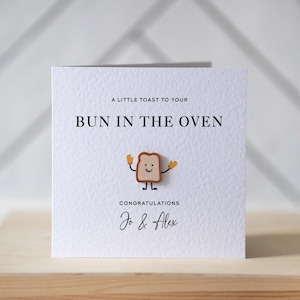 Op de afbeelding: Witte wenskaart met de tekst "A LITTLE TOAST TO YOUR BUN IN THE OVEN". Een cartoon toast karakter met een lachend gezicht en opgeheven armen staat in het midden. De kaart luidt "CONGRATULATIONS Jo & Alex."