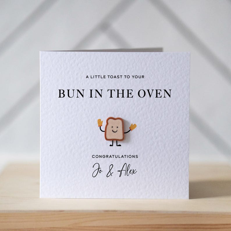 Op de afbeelding: Witte wenskaart met de tekst "A LITTLE TOAST TO YOUR BUN IN THE OVEN". Een cartoon toast karakter met een lachend gezicht en opgeheven armen staat in het midden. De kaart luidt "CONGRATULATIONS Jo & Alex."