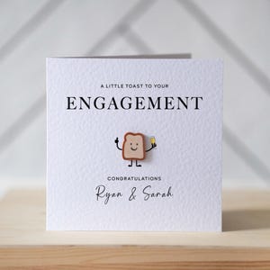 May include: White engagement card with the text "A Little Toast to Your Engagement" and "Congratulations Ryan & Sarah." A small, smiling toast character holds a champagne glass. The card is on a wooden surface.