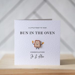 Op de afbeelding: Een witte kaart met de tekst "A LITTLE TOAST TO YOUR BUN IN THE OVEN" en "CONGRATULATIONS Jo & Alex". Een cartoon toast karakter staat in het midden. De kaart ligt op een houten oppervlak.