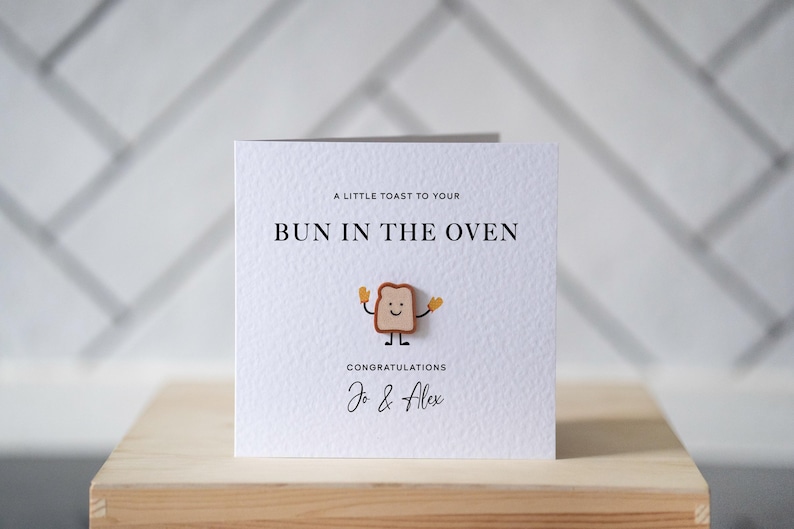 Op de afbeelding: Een witte kaart met de tekst "A LITTLE TOAST TO YOUR BUN IN THE OVEN" en "CONGRATULATIONS Jo & Alex". Een cartoon toast karakter staat in het midden. De kaart ligt op een houten oppervlak.