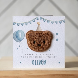 May include: A square birthday card featuring a brown teddy bear applique, a blue balloon with the number 1, and the text "HAPPY 1ST BIRTHDAY TO A BEARY SPECIAL LITTLE BOY OLIVER". The card has a white background with blue decorations.
