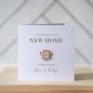 May include: A square, textured white card with the text "A LITTLE TOAST TO YOUR NEW HOME" and "CONGRATULATIONS Alex & Robyn". A cartoon toast character holds a drink. The card is on a wooden surface.