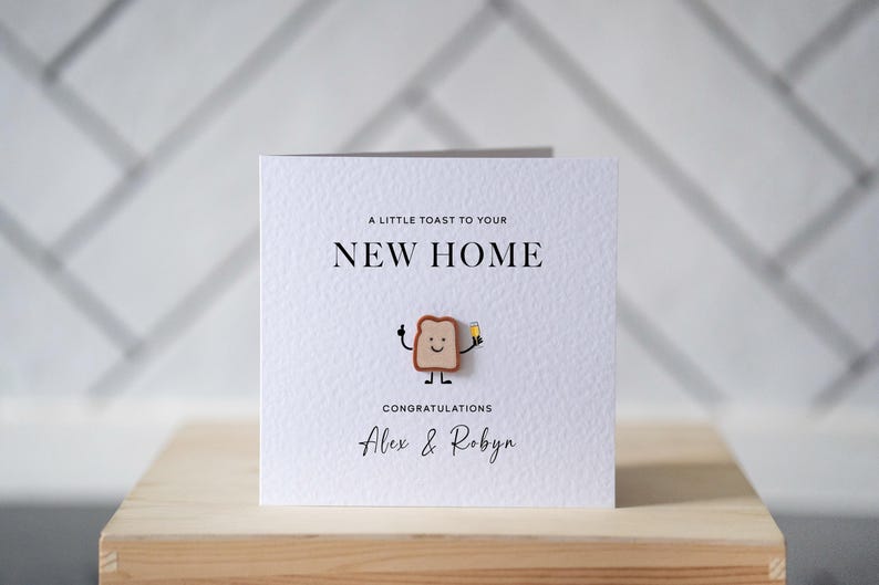 May include: A square, textured white card with the text "A LITTLE TOAST TO YOUR NEW HOME" and "CONGRATULATIONS Alex & Robyn". A cartoon toast character holds a drink. The card is on a wooden surface.