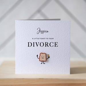 Op de afbeelding: Een witte wenskaart met de naam "Jessica" in cursief schrift. De tekst "A little toast to your DIVORCE" is vetgedrukt. Een klein, lachend toastkarakter met een drankje is aan de kaart bevestigd.