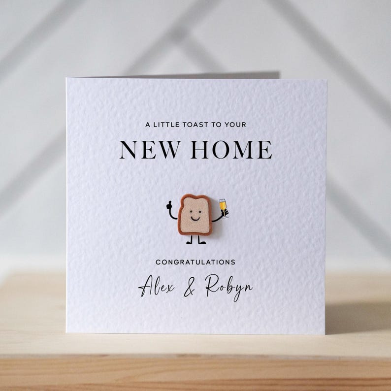 May include: A white greeting card with the text "A LITTLE TOAST TO YOUR NEW HOME" and "CONGRATULATIONS Alex & Robyn". A wooden toast character with a smiling face and a champagne glass is attached to the card.