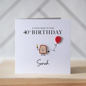 Op de afbeelding: Een witte verjaardagskaart met de tekst "A LITTLE TOAST TO YOUR 40TH BIRTHDAY" en de naam "Sarah". De kaart toont een cartoon toast karakter met een drankje en een rode ballon.