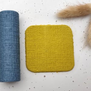 May include: A textured, cylindrical blue object and a square, yellow coaster are displayed on a speckled white surface. Two dried, fluffy, tan-coloured plant stems are also present. The objects are arranged to create a minimalist aesthetic.