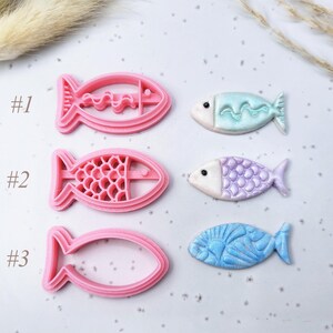 Puede incluir: Tres cortadores de galletas de plástico rosa con forma de pez y tres peces decorados en la imagen. Los peces son de color azul claro, lavanda y azul, con detalles en blanco. La imagen incluye los números #1, #2 y #3.