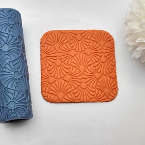 May include: A blue textured rolling pin and an orange square stamp, both with a repeating fan design. These items are likely for crafts or baking. A white flower is in the background.