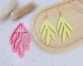 May include: A pair of pale yellow leaf-shaped earrings with gold-colored hooks rests on a wooden tray. A pink leaf-shaped cookie cutter is next to the earrings. Dried plants are in the background. The earrings and cutter are on a white surface.