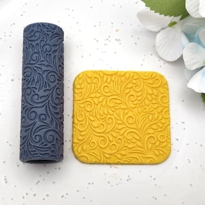 May include: A dark blue cylindrical rolling pin with an intricate embossed swirling leaf pattern next to a square piece of yellow clay featuring the same embossed design. The rolling tool and the clay square display a consistent, repeating botanical motif.