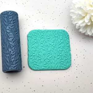 May include: A dark blue cylindrical clay roller and a square teal clay stamp, both featuring a leaf pattern. The roller is a deep blue, and the stamp is a bright teal. The leaf design is consistent on both items, suggesting they are used together.