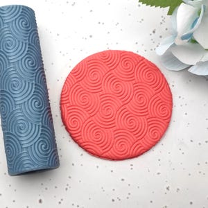 Pode incluir: Um rolo azul texturizado e um carimbo circular vermelho, ambos com um padrão em espiral. Estão expostos numa superfície branca com um detalhe floral, sugerindo um tema de artesanato ou pastelaria.